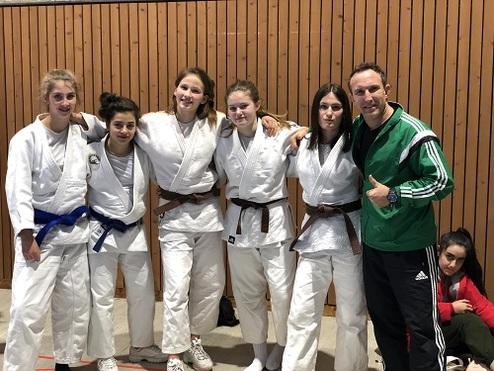 Sechs Kampfsportlerinnen in weißen Judo-Gis posieren gemeinsam mit einem Trainer in einem Sportcenter.