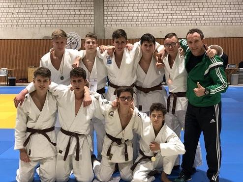 Gruppe von Judokas in Weiß mit braunen Gürteln posiert für ein Gruppenfoto auf einer blauen Judomatte.