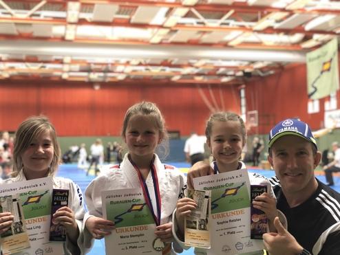 Drei Kinder im Judoanzug halten Urkunden und Medaillen, stehen lächelnd neben einem Trainer in einer Sporthalle.