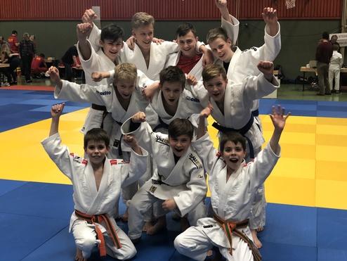 Gruppe von zehn Jungen in Judoanzügen jubelt auf einer bunten Judomatte, einige mit erhobenen Armen.