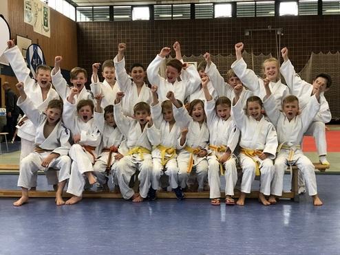 Gruppe von Kindern in weißen Judoanzügen mit gelben Gürteln, die fröhlich auf einer Bank posieren und jubeln.