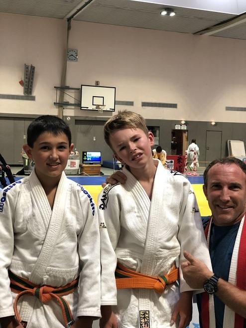 Zwei Kinder in Judogis lächeln neben einem Trainer, der freundlich auf sie zeigt, vor einem Judomattenbereich.