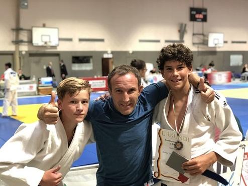 Zwei Jugendliche in Judoanzügen posieren mit einem Erwachsenen, der eine Urkunde in der Hand hält, in einer Sporthalle.