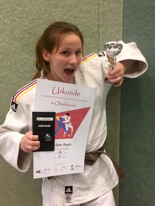 Lächelnde junge Judoka in weißem Gi hält einen Pokal und eine Urkunde mit ihrem Namen und Platzierung.