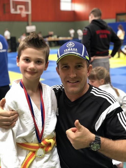 Junge mit Medaille in Judogewand neben Trainer, beide lächeln vor einer Sporthalle mit Judomatten im Hintergrund.