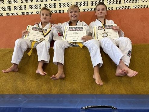 Drei Kinder in Judoanzügen halten Medaillen und Urkunden, sitzen auf einer gepolsterten Bank.