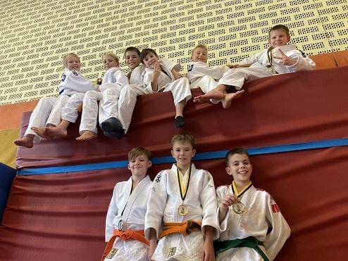 Gruppenkinder in Judoanzügen posieren auf einer Mattenwand im Trainingsraum, einige tragen Medaillen um den Hals.