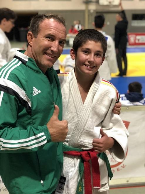 Trainer und Jugendjudoka posieren lächelnd vor einem Judomatte, beide zeigen Daumen hoch.