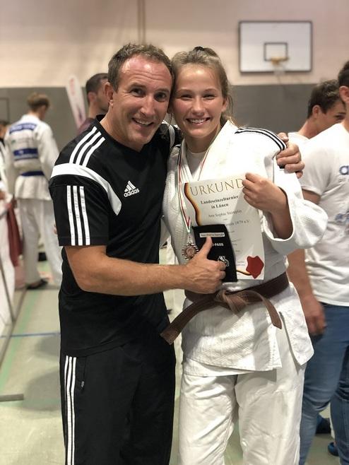 Ein Trainer umarmt eine Medaillenträgerin mit Urkunde in Judo-Gi, beide lächeln glücklich in einer Sporthalle.