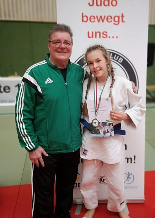 Ein Trainer steht mit einer jungen Judoka, die eine Medaille und ein Zertifikat hält, vor einem Judo-Werbebanner.