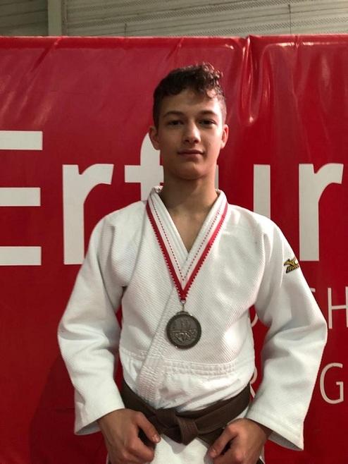 Junge in Judogi mit Silbermedaille steht vor rotem Hintergrund mit dem Schriftzug "Ertür".