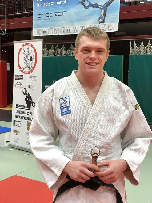 Junge im Judogi hält den Gürtel und lächelt vor einem Judoturnier-Hintergrund.