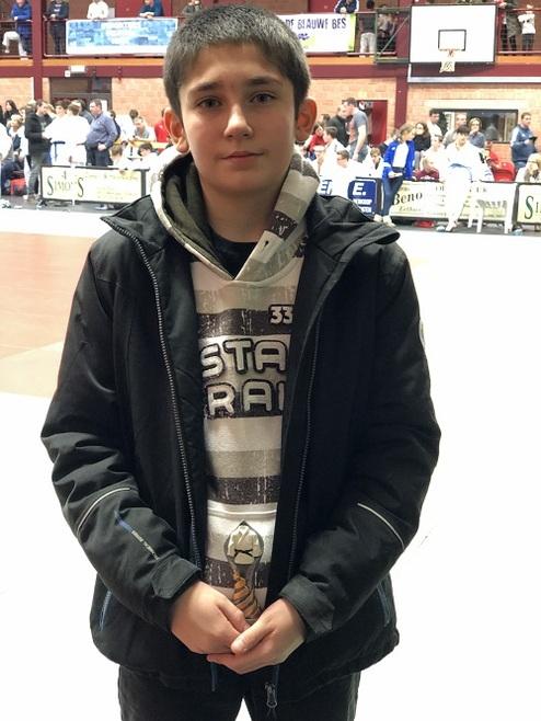 Junge mit dunklem Haar steht in Sporthalle, trägt schwarze Jacke und grauen Hoodie, im Hintergrund Zuschauer und Sportereignis.