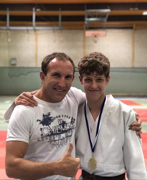 Ein Trainer mit Daumen hoch steht neben einem jugendlichen Judoka mit Medaille in einem Dojo.