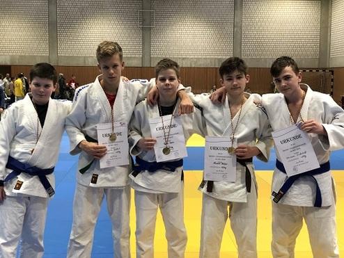 Fünf Jugendliche in Judogis halten Urkunden und Medaillen nach einem Wettkampf auf einer blauen Judomatte.