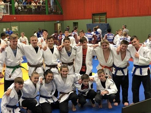 Gruppe von Jugendlichen in Judogis posiert lächelnd auf einer Judomatte während eines Turniers.