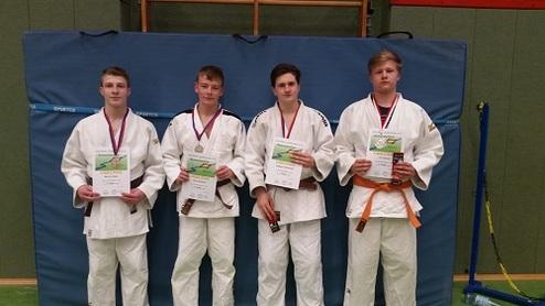 Vier Judoka in Traininganzügen halten Urkunden und Medaillen vor einer blauen Judomatte in einer Sporthalle.