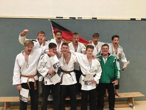 Gruppe von Judo-Athleten in weißen Kimonos mit Medaillen und Urkunden, posierend vor einer deutschen Flagge.