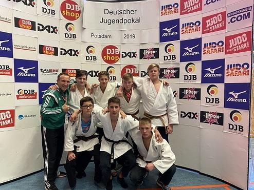 Gruppenfoto von acht Judoka in weißen Anzügen, umgeben von Sponsorenbanner beim Deutschen Jugendpokal 2019 in Potsdam.