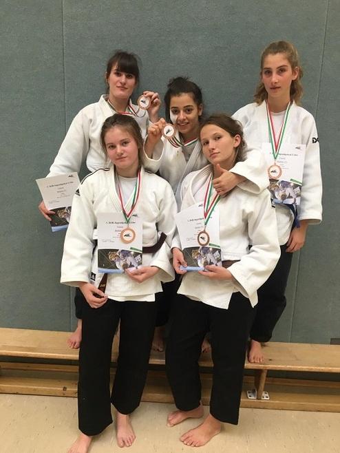 Fünf junge Judokas in weißen Anzügen halten Medaillen und Urkunden in einer Sporthalle.