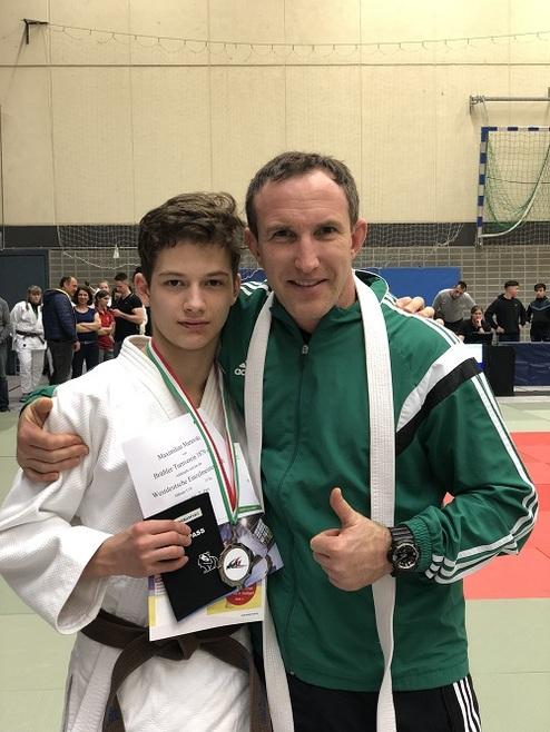 Zwei Männer in Judojacken posieren lächelnd, einer hält eine Urkunde und Medaille in der Hand.