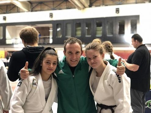 Drei Personen posieren in Judoanzügen für ein Foto in einer Sporthalle. Zwei Frauen stehen neben einem Trainer.