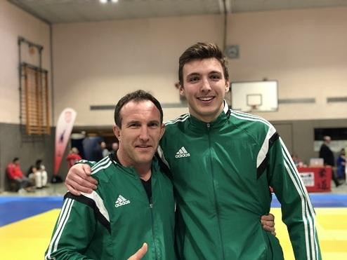 Zwei Männer in grünen Sportjacken stehen lächelnd nebeneinander auf einer Judo-Matte in einer Sporthalle.