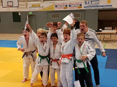 Gruppe junger Judoka in weißen Kimonos jubelt mit Medaillen und einem Pokal nach einem Wettkampf auf der Judomatte.