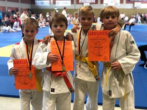 Vier Jungen in Judoanzügen halten stolz Urkunden und Medaillen in einer Turnhalle während eines Wettbewerbs.