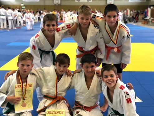 Sechs Judo-Jungs in weißen Anzügen posieren lächelnd auf einer blauen Matte, zwei Halten Urkunden in der Hand.