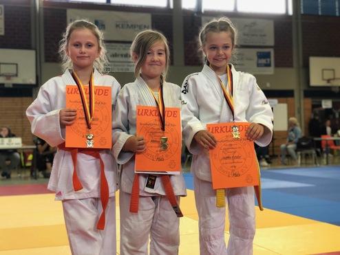 Drei junge Judokas in weißen Judogis halten Urkunden und Medaillen nach einem Wettkampf in einer Sporthalle.