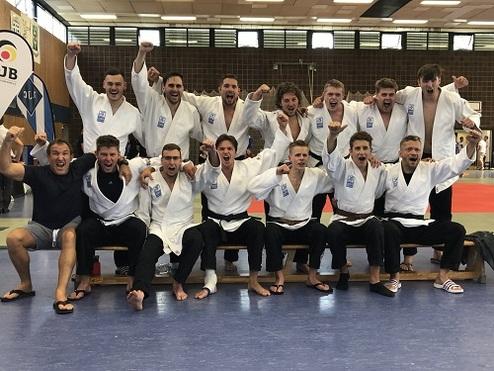 Gruppe von 14 Judo-Kämpfern in weißen Anzügen, jubelnd auf einer Tribüne, in einer Sporthalle.