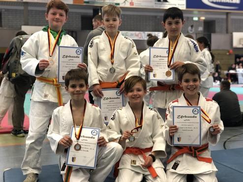 Sechs Kinder in Judogis posieren stolz mit Medaillen und Urkunden nach einem Wettkampf. Hinter ihnen ist eine Sporthalle sichtbar.
