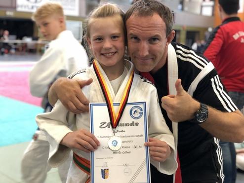 Mädchen in Judogewand hält Siegerurkunde und Medaille, steht mit Trainer in einer Sporthalle.