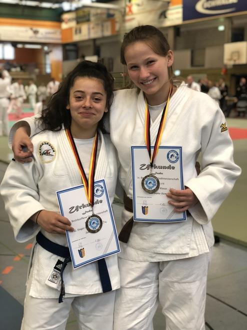 Zwei Judokas in Uniform halten stolz ihre Urkunden und Medaillen nach einem Wettkampf in der Halle.
