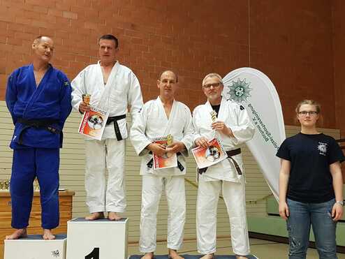 Vier Judoka in Gi stehen auf Podeststufen, jeweils mit Trophäen und Urkunden, während ein Mädchen daneben beobachtet.