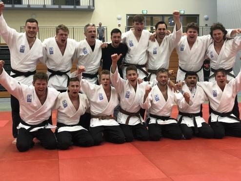Gruppe von Judo-Kämpfern in weißen Judogis posiert freudig auf einer Judo-Matte und jubelt gemeinsam.