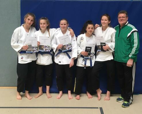 Fünf Judokas in Weiß mit blauen Gürteln halten Urkunden, zusammen mit einem Trainer in einem Sportraum.