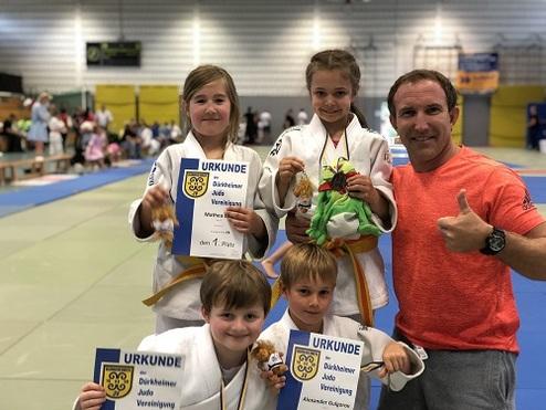 Vier Kinder in Judoanzügen halten Urkunden und Medaillen, stehen in einer Turnhalle neben einem Erwachsenen, der Daumen hoch zeigt.