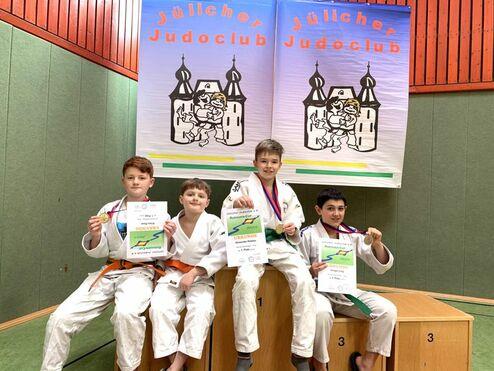 Vier Kinder in Judokleidung sitzen auf einer Treppe, halten Medaillen und Urkunden, mit Bannern des Jülicher Judoclubs im Hintergrund.