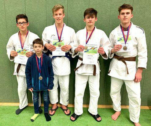 Vier Judoka in weißen Anzügen mit Medaillen und Urkunden stehen vor einer grünen Wand; ein Junge in blauer Jacke in der Mitte.