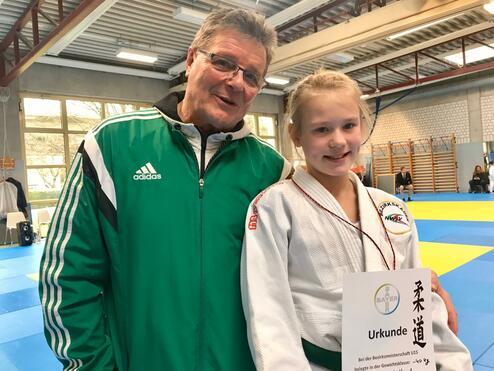 Lächelnder Trainer mit einem jungen Mädchen in Judomantel, das stolz eine Urkunde in der Halle hält.