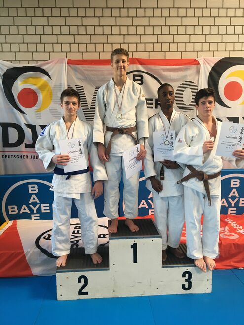 Vier Judoka stehen auf dem Siegertreppchen, halten Urkunden und Medaillen, mit einem Hintergrund von Judoverband-Logos.