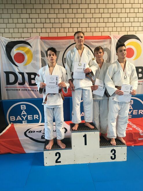 Vier Jugendliche in Judojacken stehen auf einem Podest und halten Urkunden. Im Hintergrund sind Wettbewerbsbanner zu sehen.
