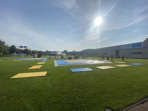 Veranstaltungsfläche mit gelben und blauen Matten auf grüner Wiese, im Hintergrund Gebäude und Personen bei Aktivitäten.