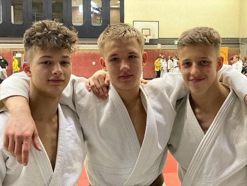 Drei Jugendliche in weißen Judoanzügen stehen mit aufeinanderliegenden Armen in einer Sporthalle.