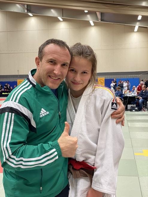 Ein Mann und ein Mädchen in Judokleidung posieren lächelnd vor einer Wettkampfschule mit Zuschauern im Hintergrund.