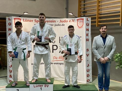Drei Judoka stehen auf einem Siegertreppchen mit Urkunden, umgeben von einer Bannerwand des Lüner SV.