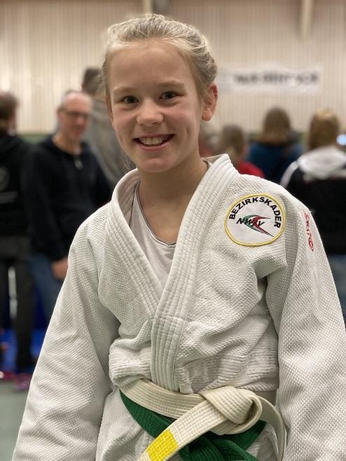 Lächelndes Mädchen in weißem Judogi mit grünem Gürtel, steht vor einer Menschenmenge in einer Sporthalle.