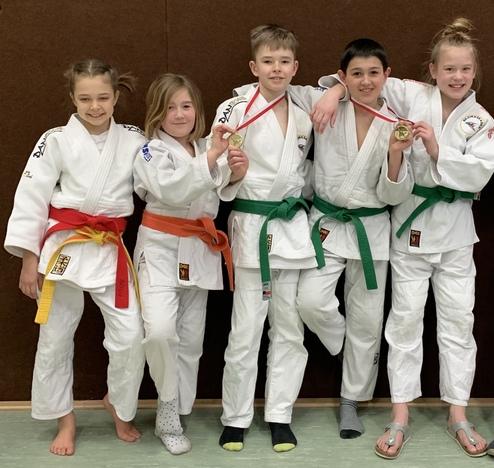 Fünf Kinder in Judoanzügen mit verschiedenen Gürtelfarben posieren stolz mit Medaillen vor einer braunen Wand.